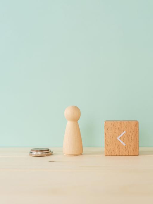 Zwei Spielfiguren aus Holz stehen auf einem Tisch. Dazwischen ein Holzklötzchen, auf dem das 
Größer-als-Zeichen zu sehen ist, es zeigt nach links. Neben der linken Figur stapeln sich vier Geldstücke, neben der rechten Figur deutlich mehr.

