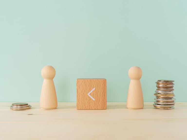 Zwei Spielfiguren aus Holz stehen auf einem Tisch. Dazwischen ein Holzklötzchen, auf dem das 
Größer-als-Zeichen zu sehen ist, es zeigt nach links. Neben der linken Figur stapeln sich vier Geldstücke, neben der rechten Figur deutlich mehr.
