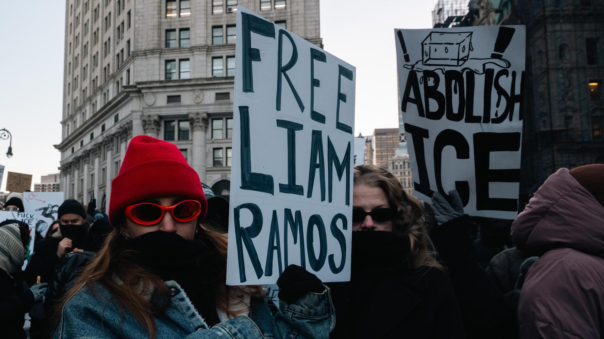 Die Frau mit roter Mütze, Sonnenbrille und Schal über dem Mund hält ein Pappschild mit der Aufschrift "Free Liam Ramos". Daneben weitere Demonstranten, einer hält ein Schild mit der Aufschrift "Abolish ICE". 