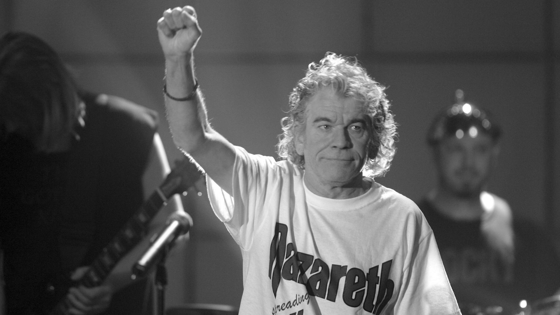 Auf dem schwarz-weiß-Bild steht McCafferty mit einem hellen Nazareth-Shirt auf der Bühne und reckt den rechten Arm hoch. Er lächelt leicht. Dahinter zwei weitere Personen.