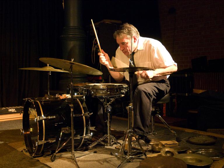 Ein Mann mit weißem Hemd und schwarzer Krawatte sitzt in charakteristisch gebeugter Haltung am Drumset und spielt
