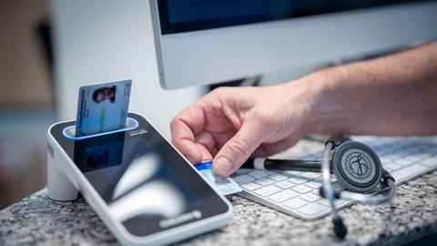 Eine Hand steckt den Arztausweis in ein Lesegerät für die elektronische Patientenakte (ePA). Auch eine Krankenkassenkarte wird eingesteckt und ausgelesen.