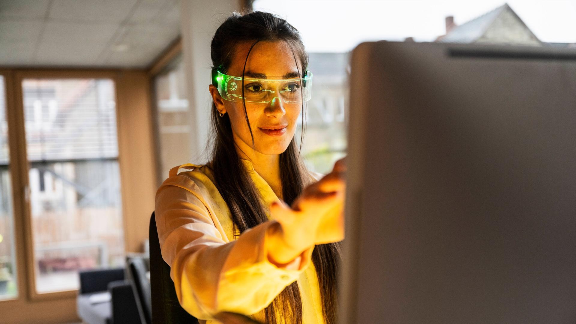 Eine Frau hat eine Smartbrille auf und sitzt vor einem PC.