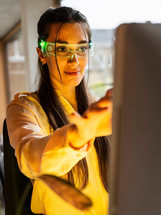 Eine Frau hat eine Smartbrille auf und sitzt vor einem PC.