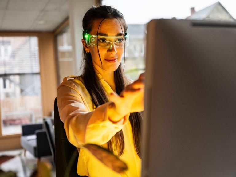 Eine Frau hat eine Smartbrille auf und sitzt vor einem PC.