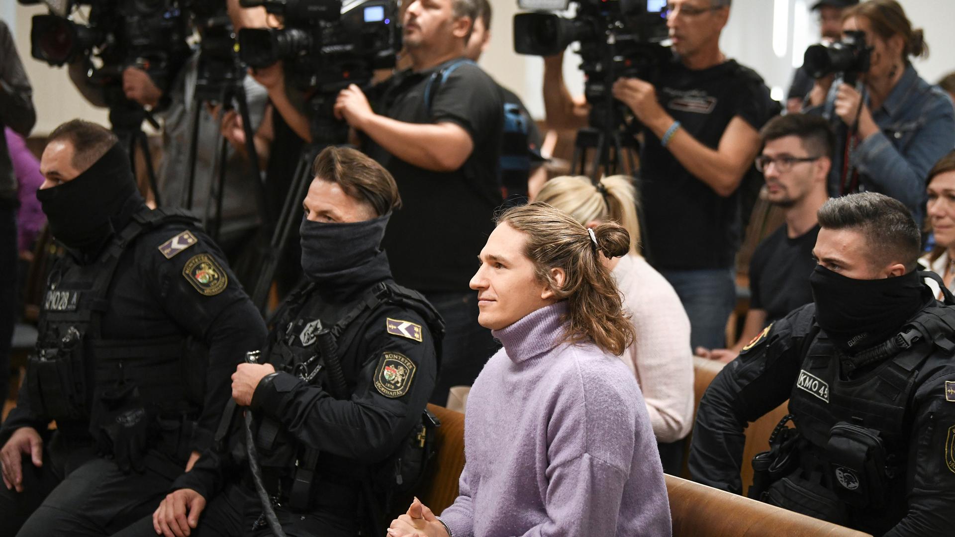 Maja T. sitzt auf der Anklagebank in Budapest. Um sie herum zahlreiche in schwarz gekleidete Polizisten, dahinter mehrere Kameramänner. Maja T. sitzt auf der Anklagebank in Budapest. Um sie herum zahlreiche in schwarz gekleidete Polizisten, dahinter mehrere Kameramänner.