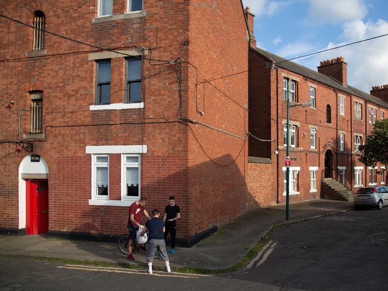 Jugendliche stehen zusammen auf einer Straße vor einer Häuserreihe ehemaliger Arbeitersiedlungen in Dublin. 