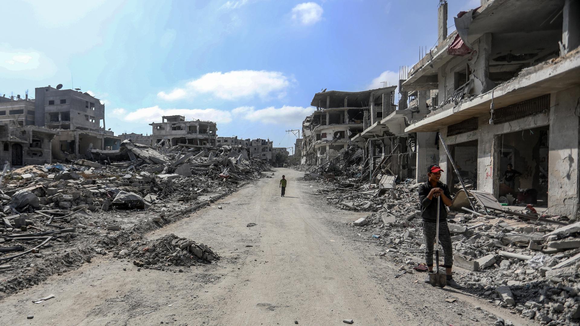 Eine Straße in der Stadt Chan Junis im Gazastreifen. Zahlreiche Gebäude sind in Trümmern. Eine Frau steht mit einer Schaufel am Rand.