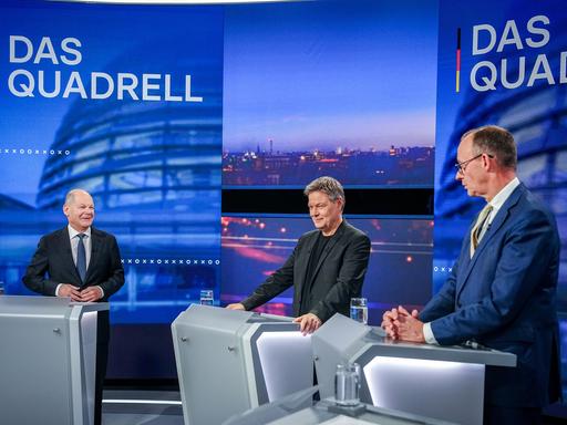 Drei Männer in Anzügen stehen an Rednerpulten in einem Fernsehstudio.