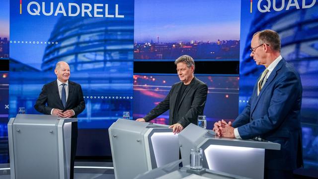 Drei Männer in Anzügen stehen an Rednerpulten in einem Fernsehstudio.