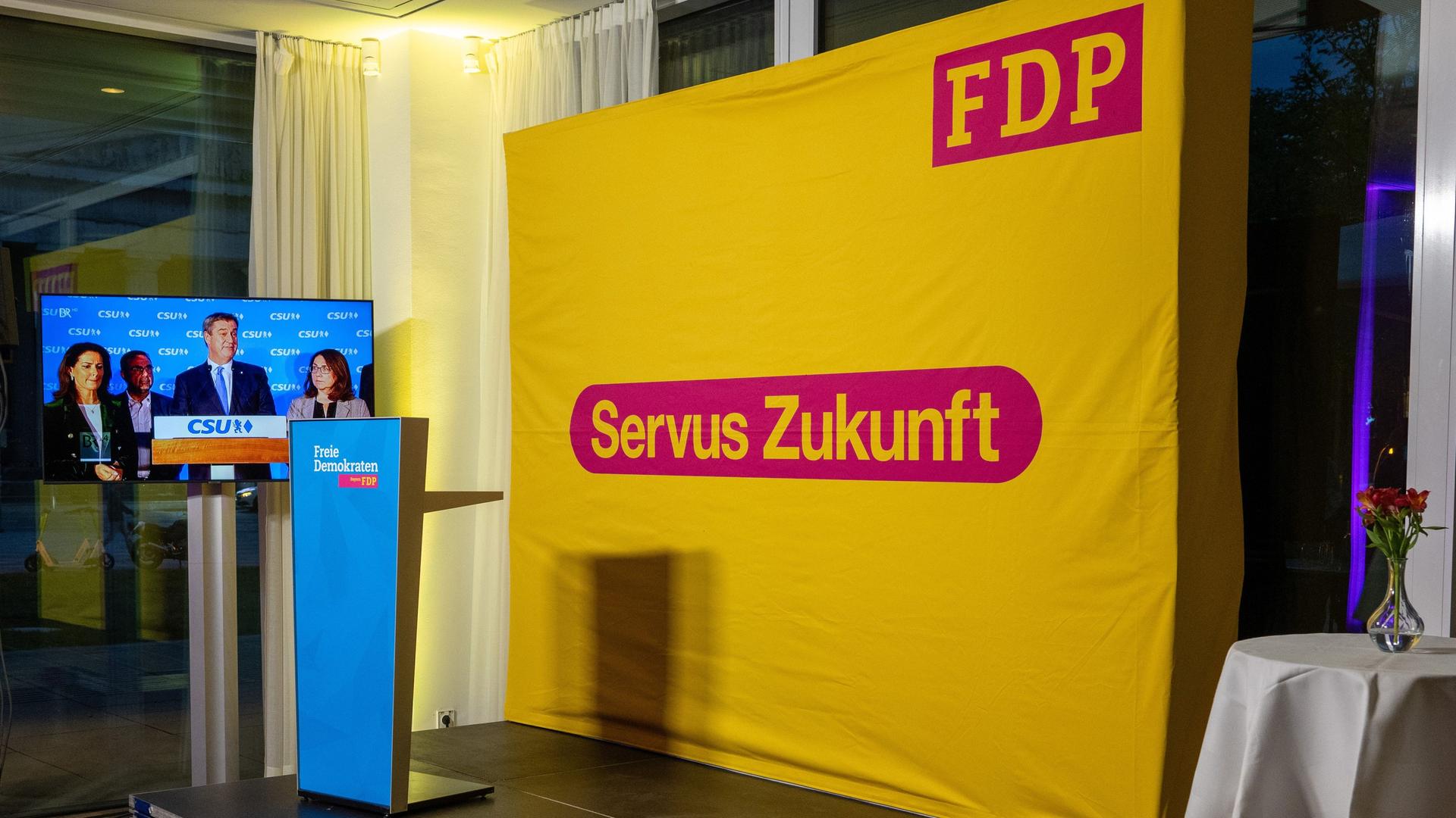 Markus Söder (CSU), Ministerpräsident von Bayern, ist bei bei der Wahlparty der FDP auf einem Bildschirm hinter leeren Stehtischen zu sehen. Auf einem Großplakat steht "Servus Zukunft - FDP"
