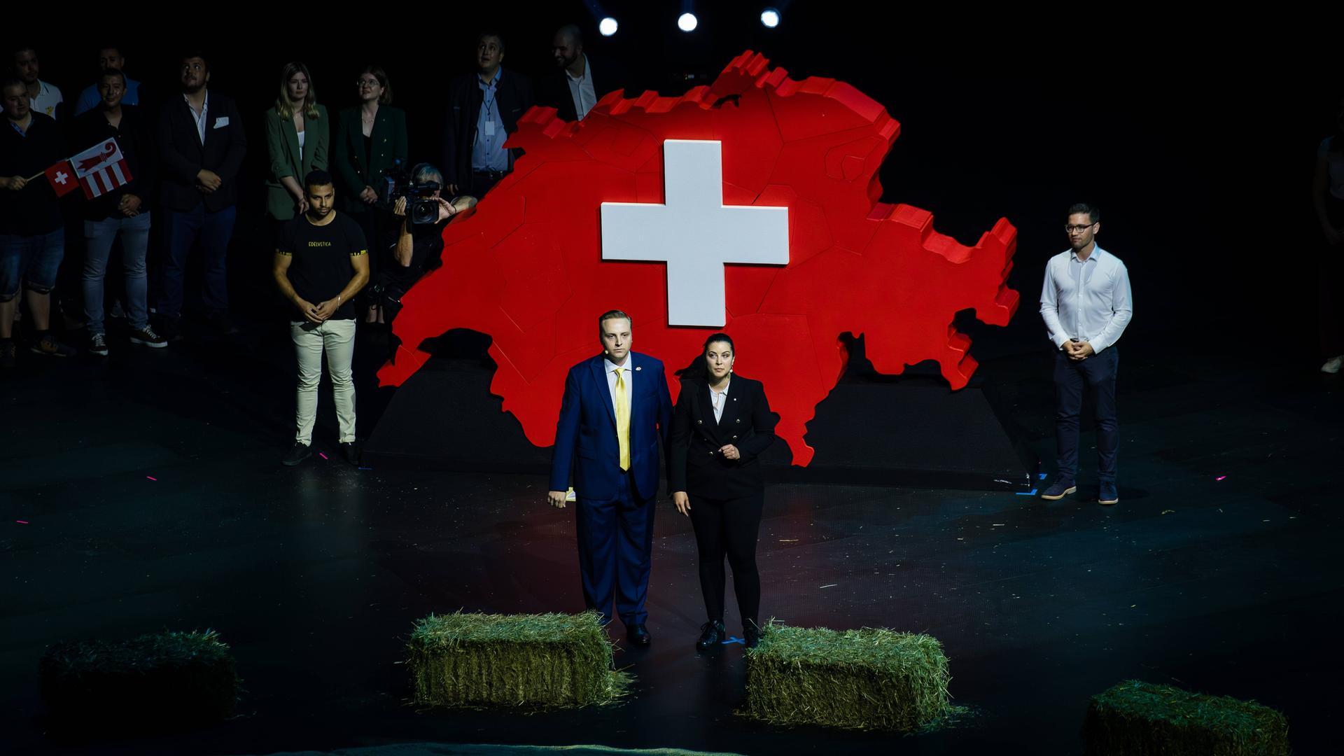 Ein Mann und eine Frau stehen auf einer Bühne, die durch Heuballen begrenzt wird. Hinter ihnen prangt ein großer roter Umriss der Schweiz mit einem weißen Kreuz in der Mitte.