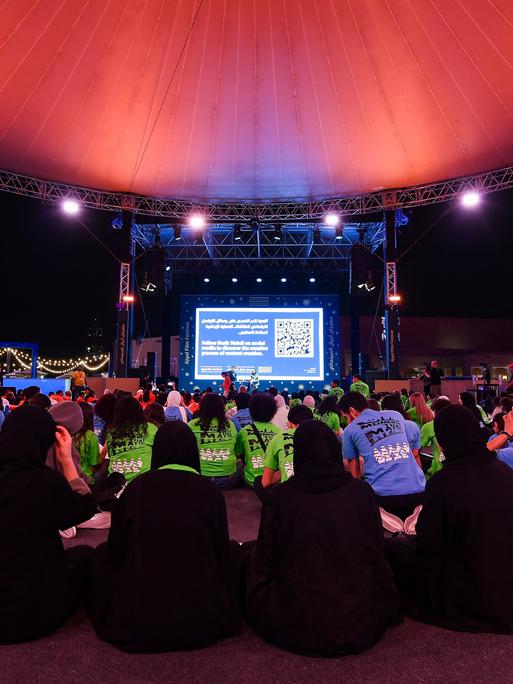 Die Eröffnung des Ajyal Film Festivals 2024 in Katar. Die Besucher sitzen auf dem Boden, einige Frauen verhüllt, andere in westlicher Kleidung. 
