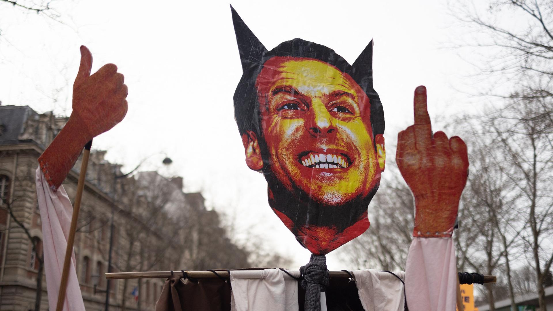 Protestler halten ein Schild hoch, das Frankreichs Präsident Macron mit Teufelshörnern und ausgestrecktem Mittelfinger zeigt