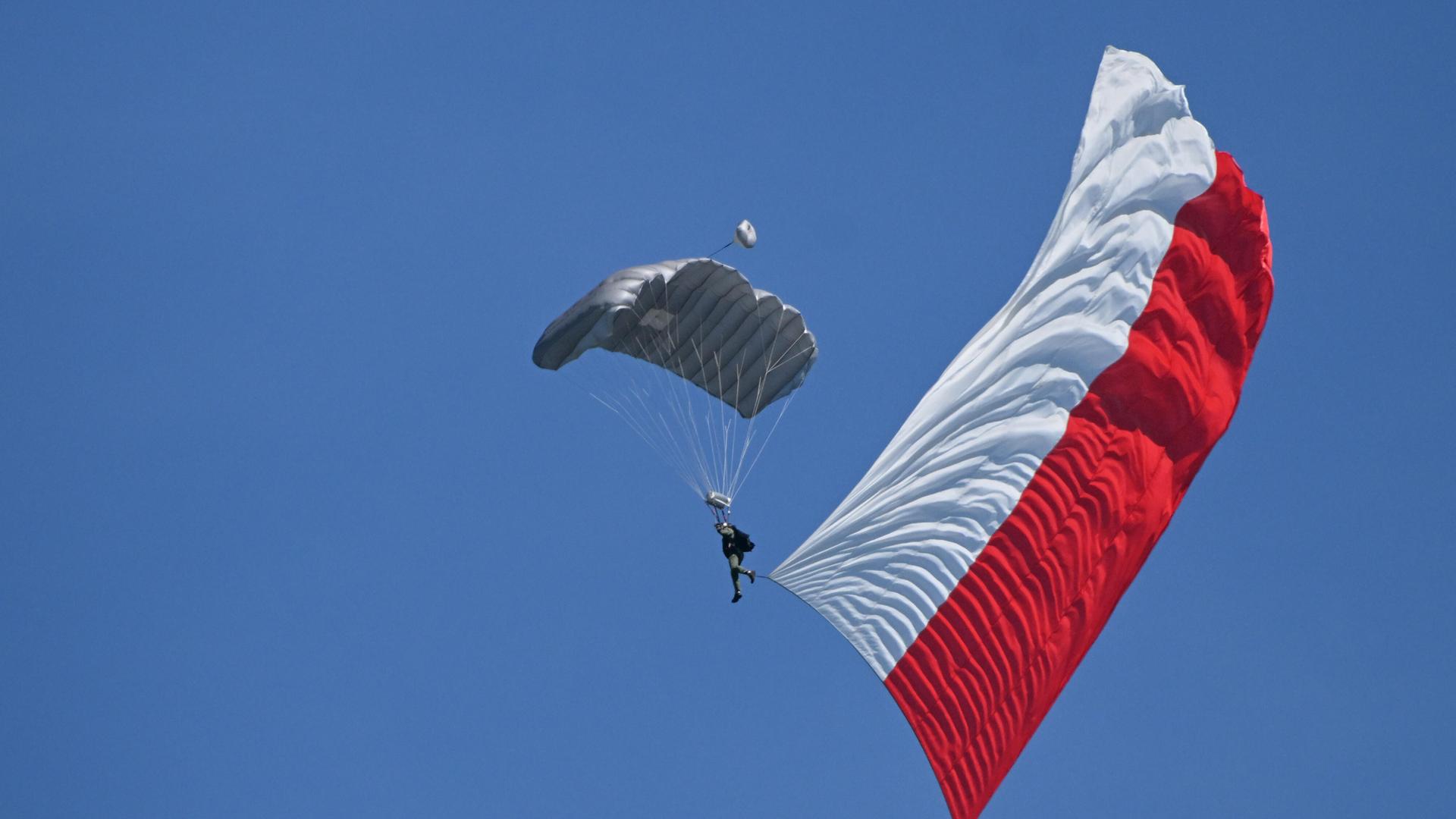 Zu sehen ist ein Fallschirmspringer, der in der Luft die polnische Flagge hinter sich ausbreitet. Der Himmel ist wolkenlos blau.