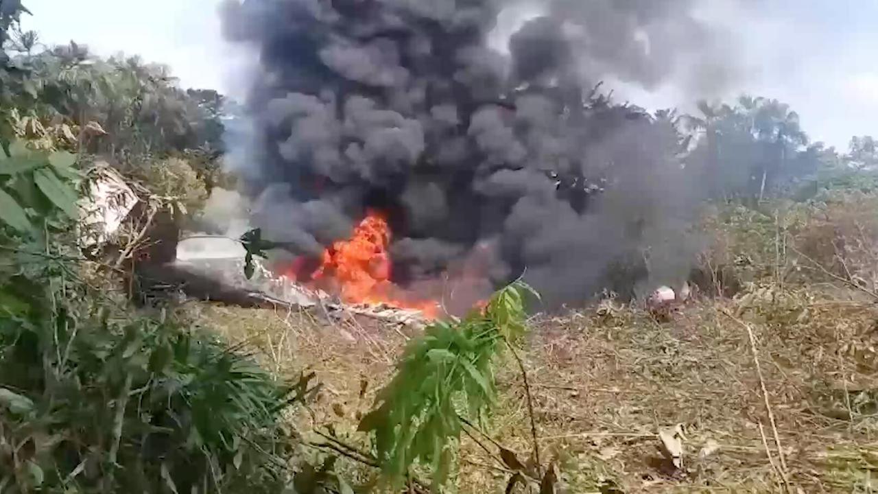 Das Bild zeigt das brennende Wrack in einer urwaldähnlichen Umgebung, aus dem dichter schwarzer Rauch aufsteigt. 