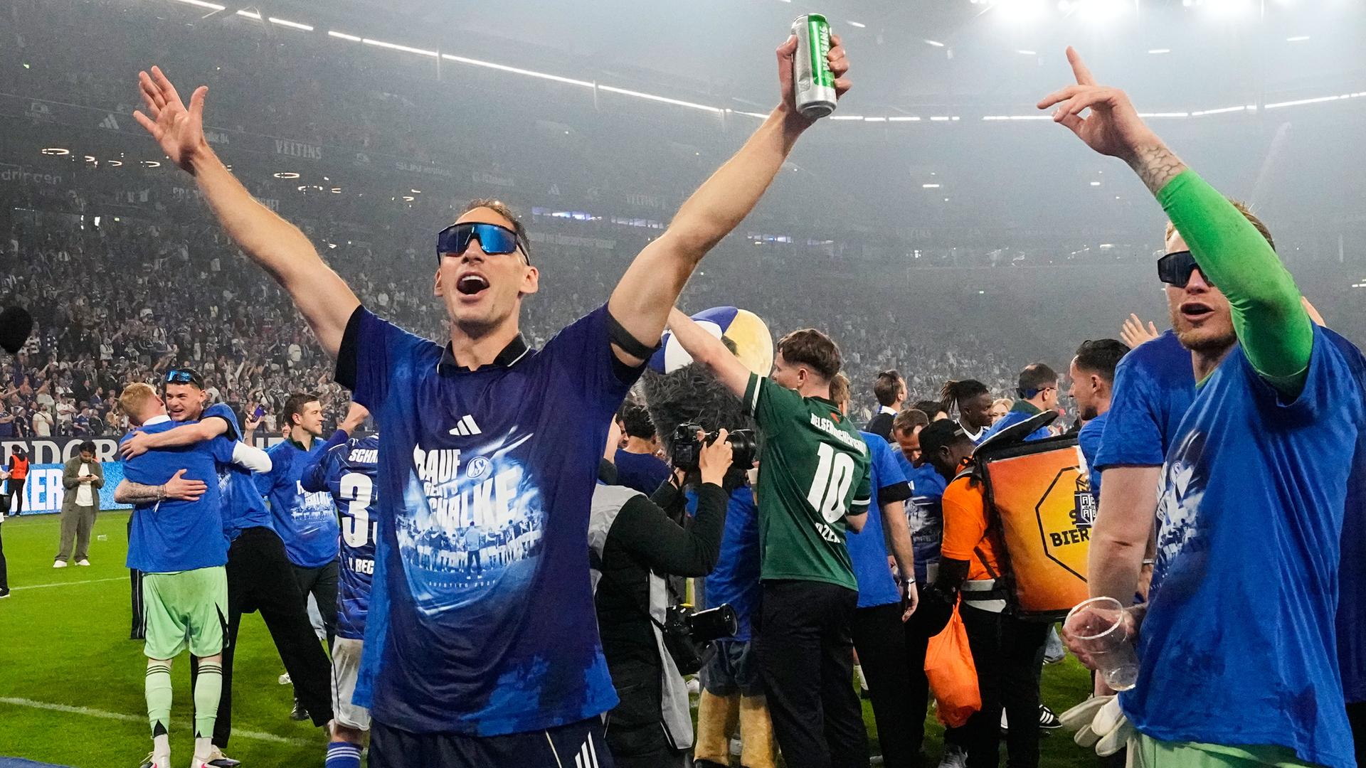 Schalkes Timo Becker und Schalkes Torwart Loris Karius (v.l.) mit Sonnenbrillen und Becker mit Bierdose in der Hand jubeln. Im Hintergrund feiern Spieler und Trainerteam im Stadion.