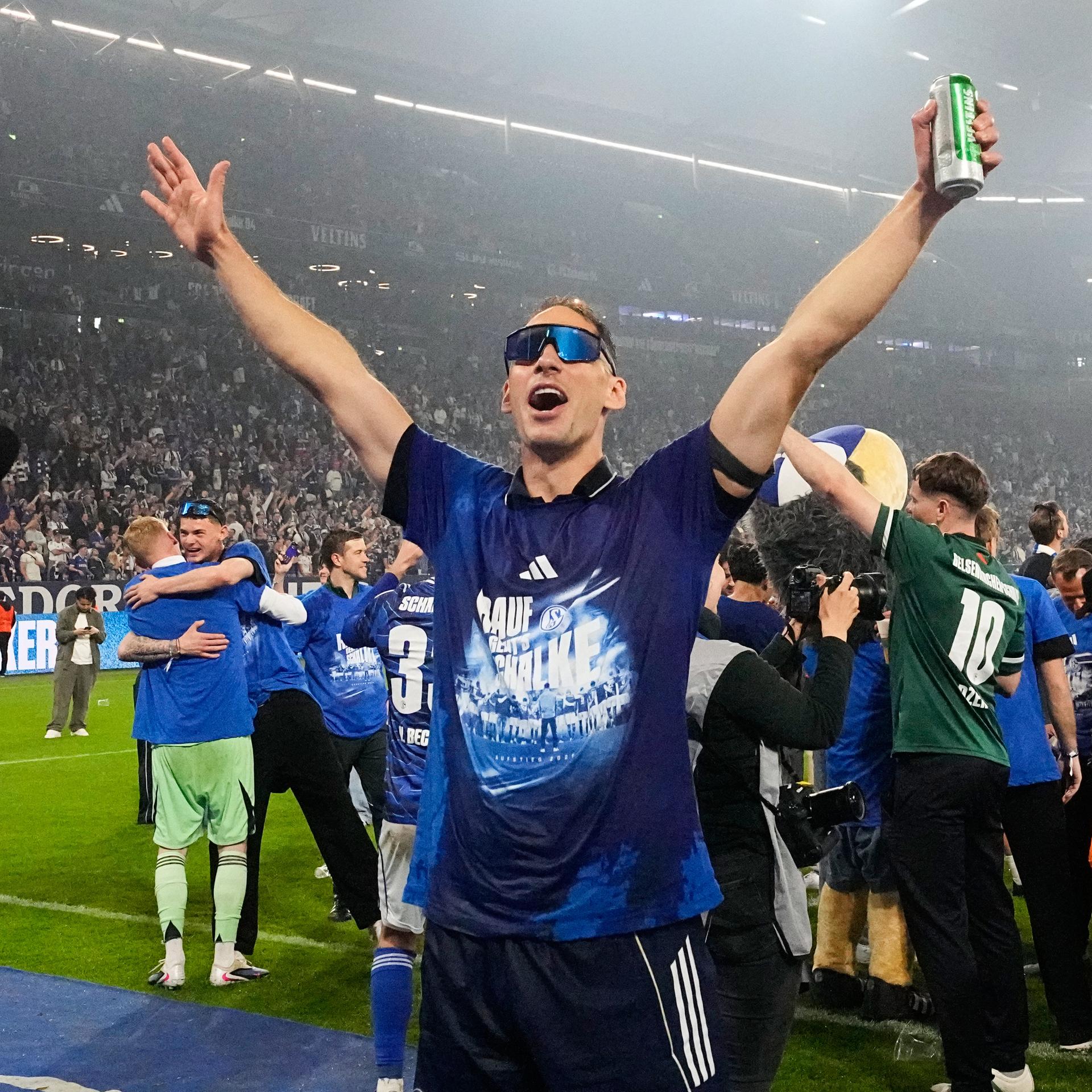 Schalkes Timo Becker und Schalkes Torwart Loris Karius (v.l.) mit Sonnenbrillen und Becker mit Bierdose in der Hand jubeln. Im Hintergrund feiern Spieler und Trainerteam im Stadion.