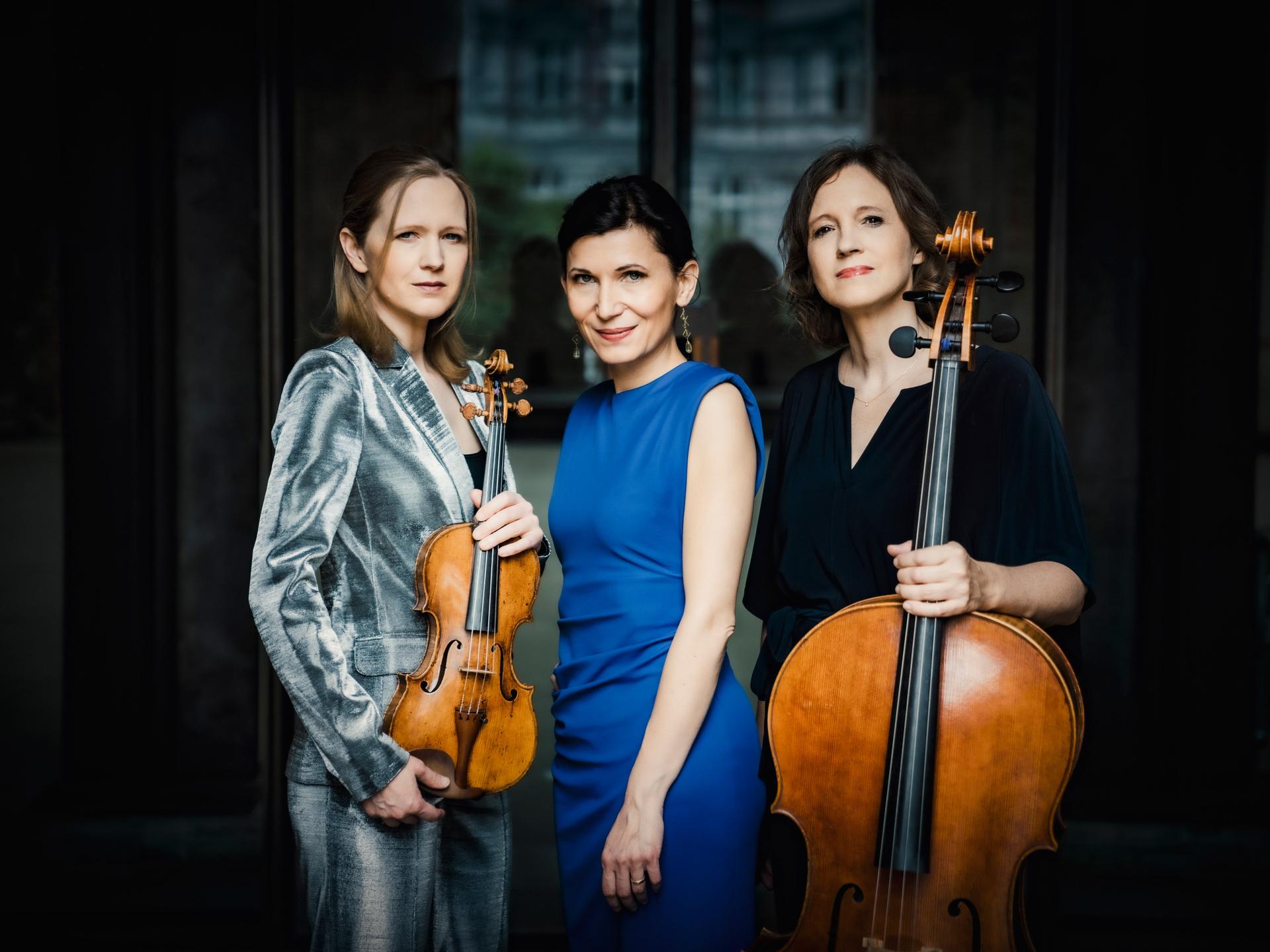 Drei Musikerinnen vor dunklem Hintergrund. Links, in einem silbernen Samtanzug, die Geigerin, die Pianistin in blauem Kleid in der Mitte und rechts eine dunkelhaarige Cellistin mit ihrem Instrument in der Hand. Alle drei schauen in die Kamera.