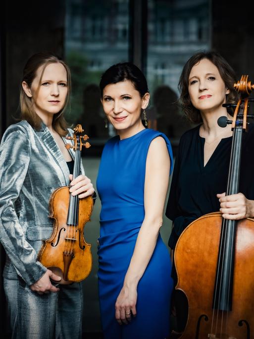 Drei Musikerinnen vor dunklem Hintergrund. Links, in einem silbernen Samtanzug, die Geigerin, die Pianistin in blauem Kleid in der Mitte und rechts eine dunkelhaarige Cellistin mit ihrem Instrument in der Hand. Alle drei schauen in die Kamera.