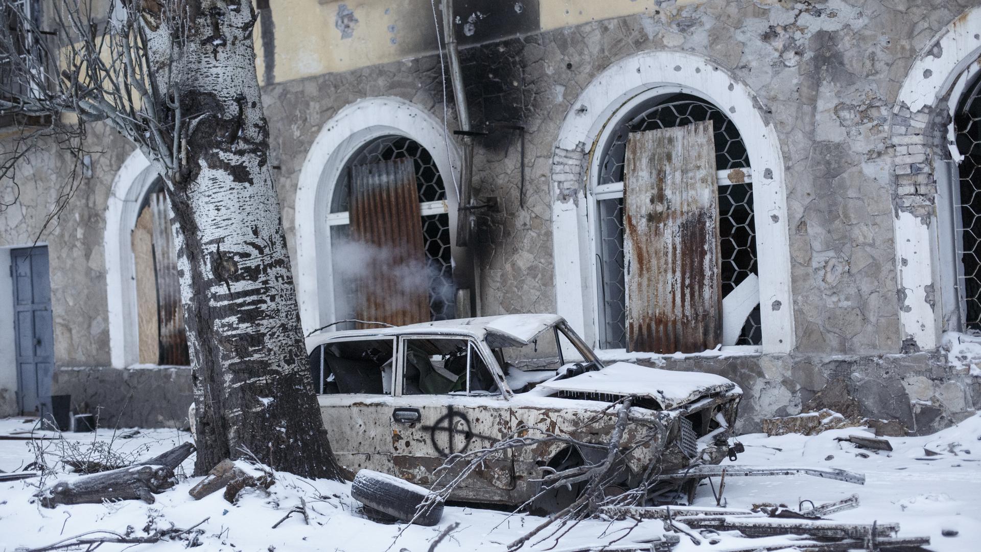 Ein zerstörtes Fahrzeug steht vor einem zerstörten Gebäude in der Ukraine, es liegt Schnee.