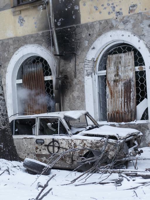 Ein zerstörtes Fahrzeug steht vor einem zerstörten Gebäude in der Ukraine, es liegt Schnee.