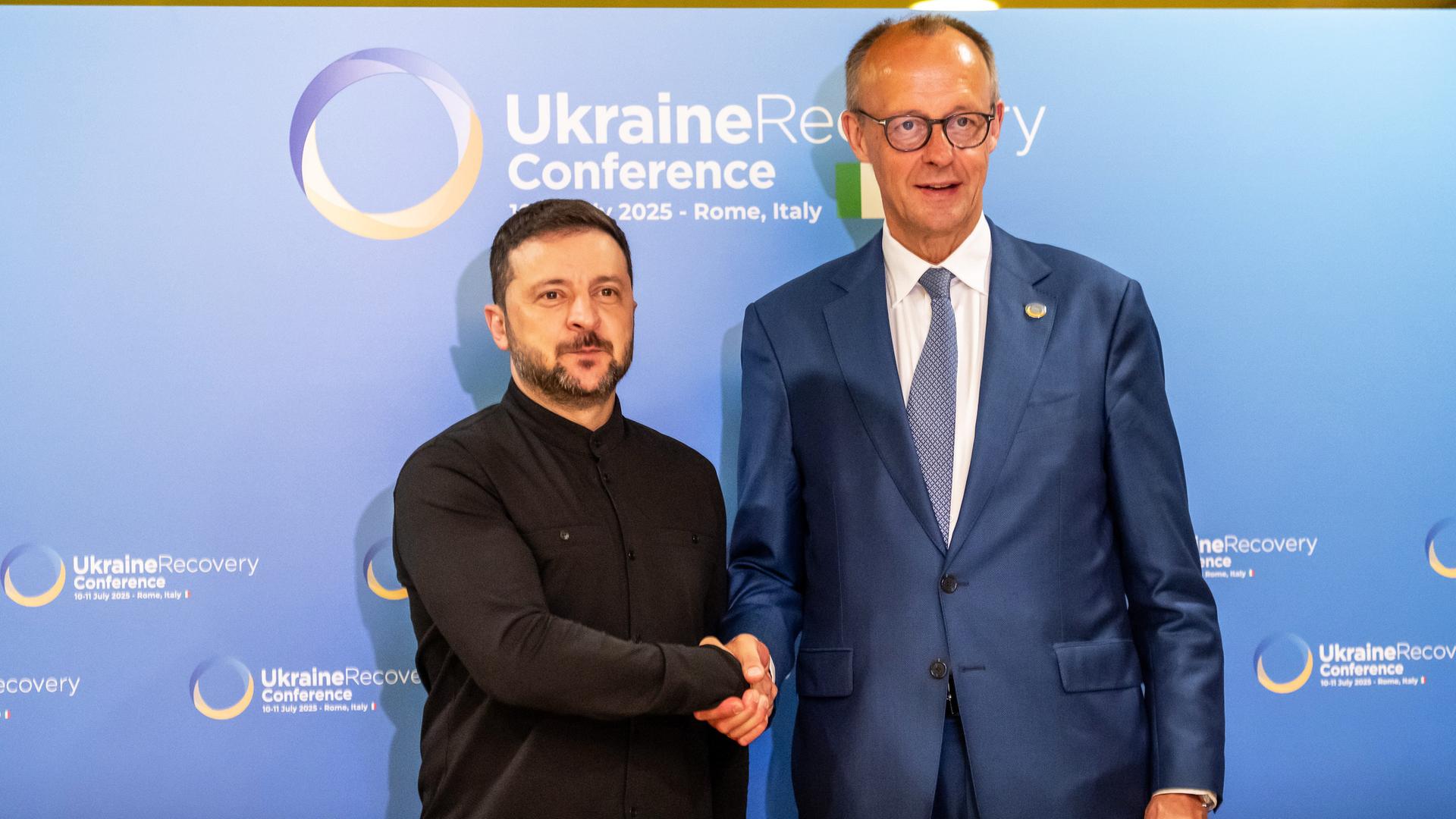 Friedrich Merz und Wolodymyr Selenskyj geben sich die Hand. Im Hintergrund ist auf einer Stellwand das Logo der Wiederaufbaukonferenz für die Ukraine zu sehen.