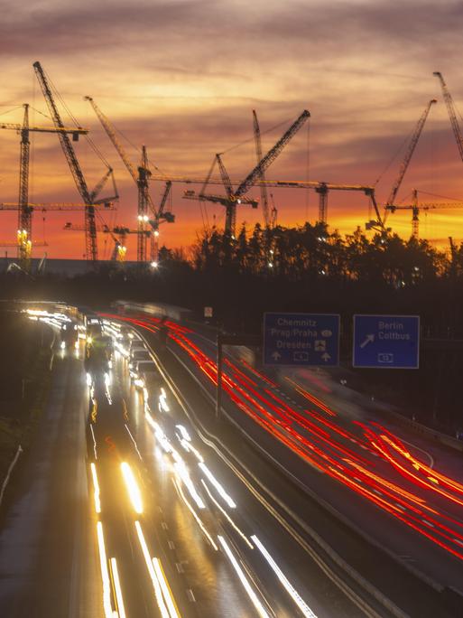 Baustelle der ESMC-Chipfabrik: Daran vorbei fahren in der Dämmerung Autos auf der Autobahn. Die Lichter sind als weiße und rote Schweife zu erkennen. Im Hintergrund stehen Krähne der Baustelle. Baustelle der ESMC-Chipfabrik: Daran vorbei fahren in der Dämmerung Autos auf der Autobahn. Die Lichter sind als weiße und rote Schweife zu erkennen. Im Hintergrund stehen Krähne der Baustelle.