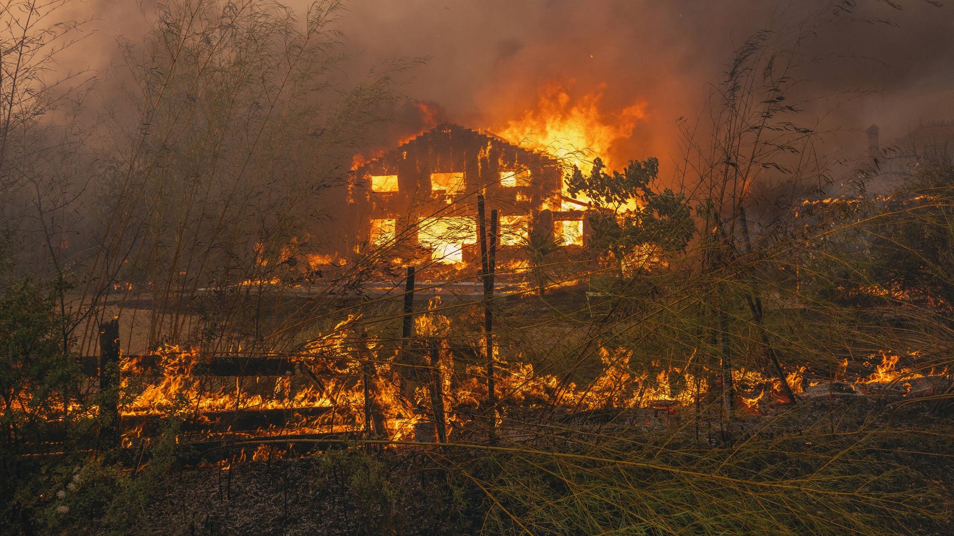Blick aus der Ferne: Ein Haus und ein Zaun davor brennen lichterloh. Alles ist rot und verraucht.