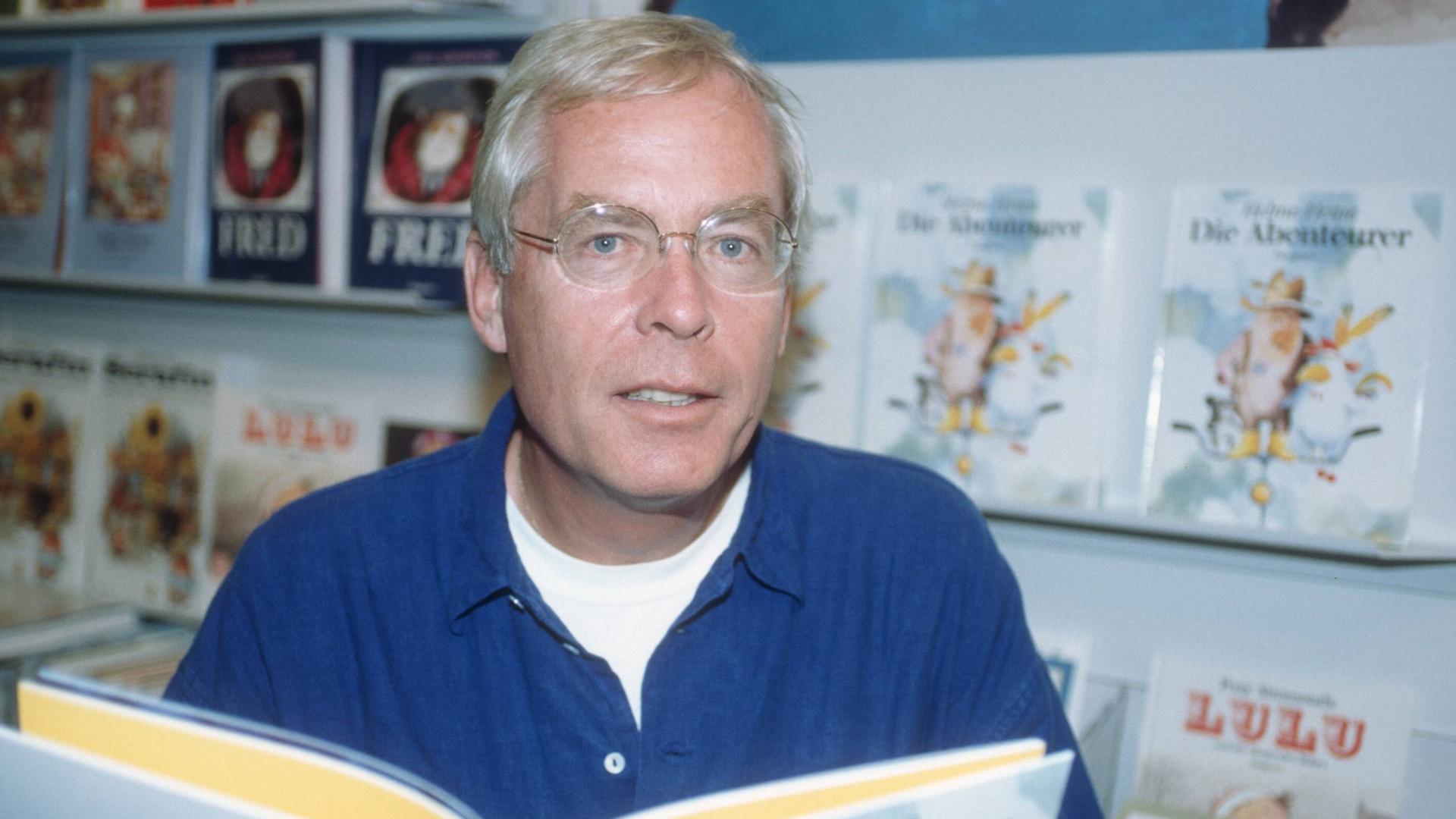 Helme Heine blickt vor Buchregalen in die Kamera und hält ein aufgeschlagenes Buch vor sich Helme Heine blickt vor Buchregalen in die Kamera und hält ein aufgeschlagenes Buch vor sich