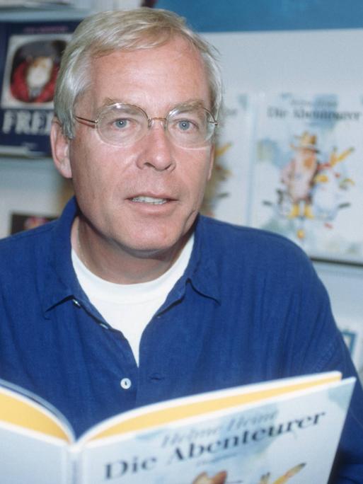 Helme Heine blickt vor Buchregalen in die Kamera und hält ein aufgeschlagenes Buch vor sich