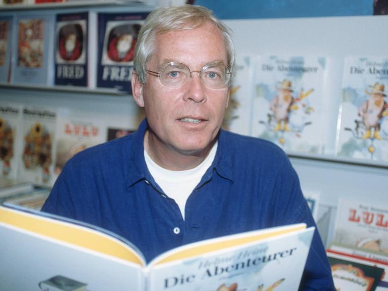 Helme Heine blickt vor Buchregalen in die Kamera und hält ein aufgeschlagenes Buch vor sich