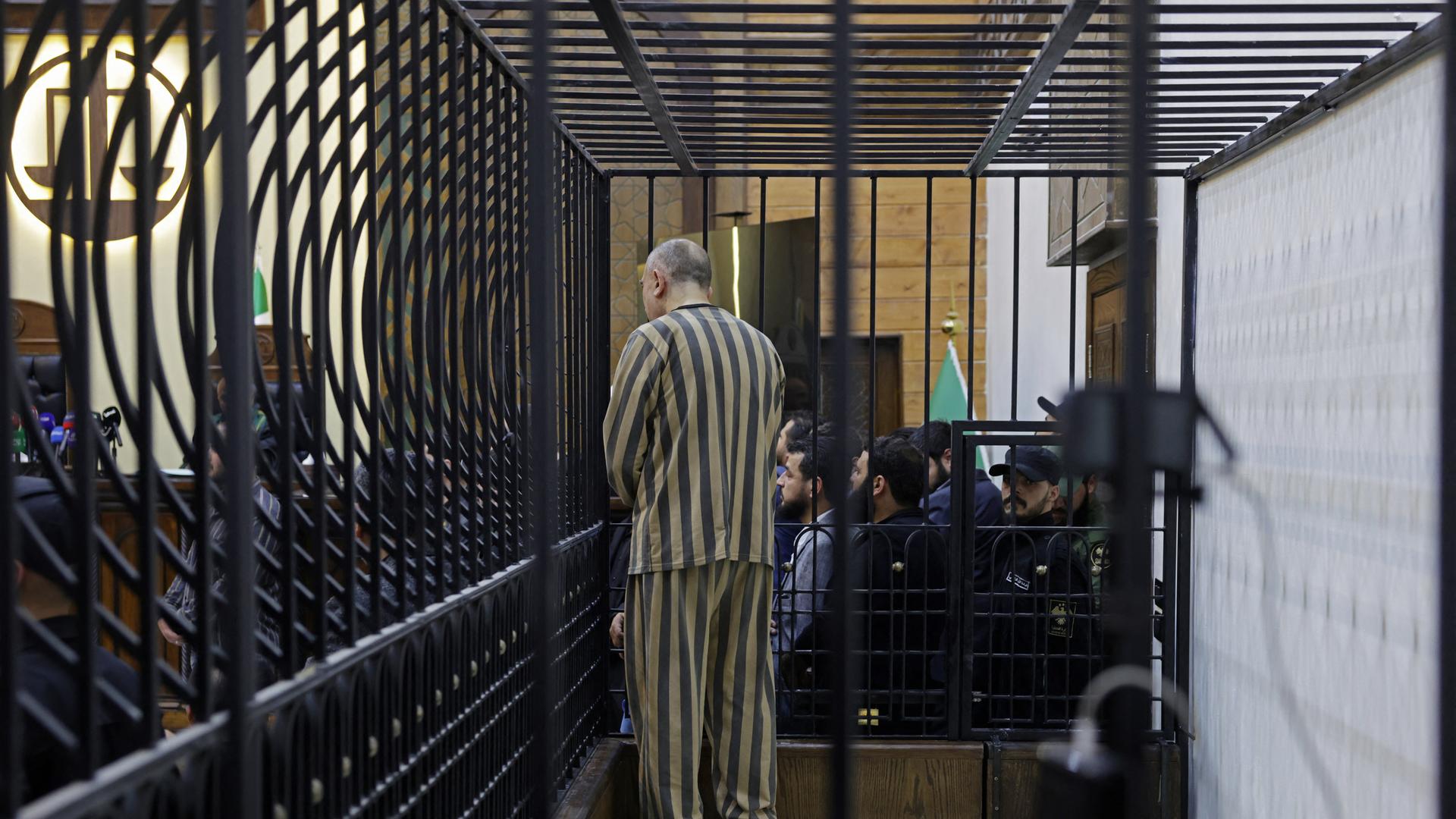 Atif Nadschib wird im Justizpalast in Damaskus der Prozess gemacht. Er trägt gestreifte Häftlingskleidung und steht in einer Kammer hinter Gittern. 