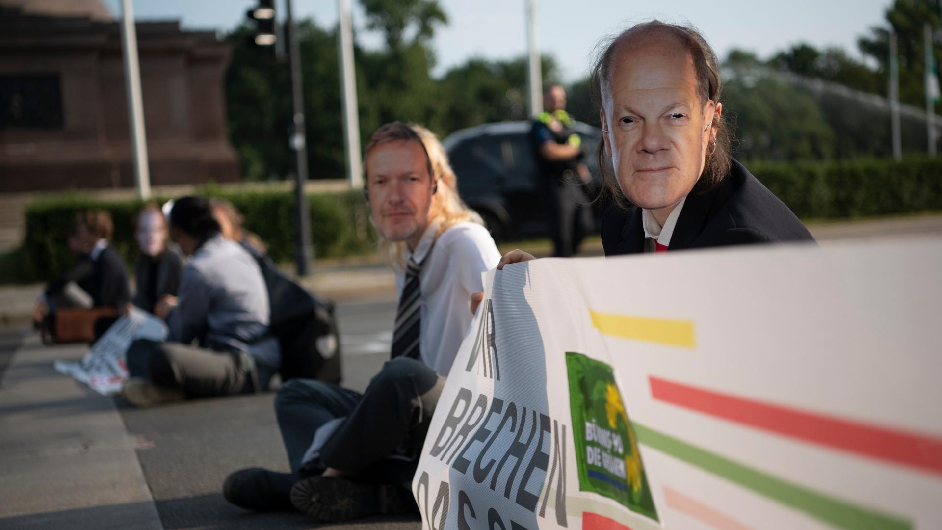 Zwei Demonstranten sitzen auf der Straße. Sie tragen Masken mit Fotos von Bundeskanzler Scholz und Bundeswirtschaftsminister Habeck.