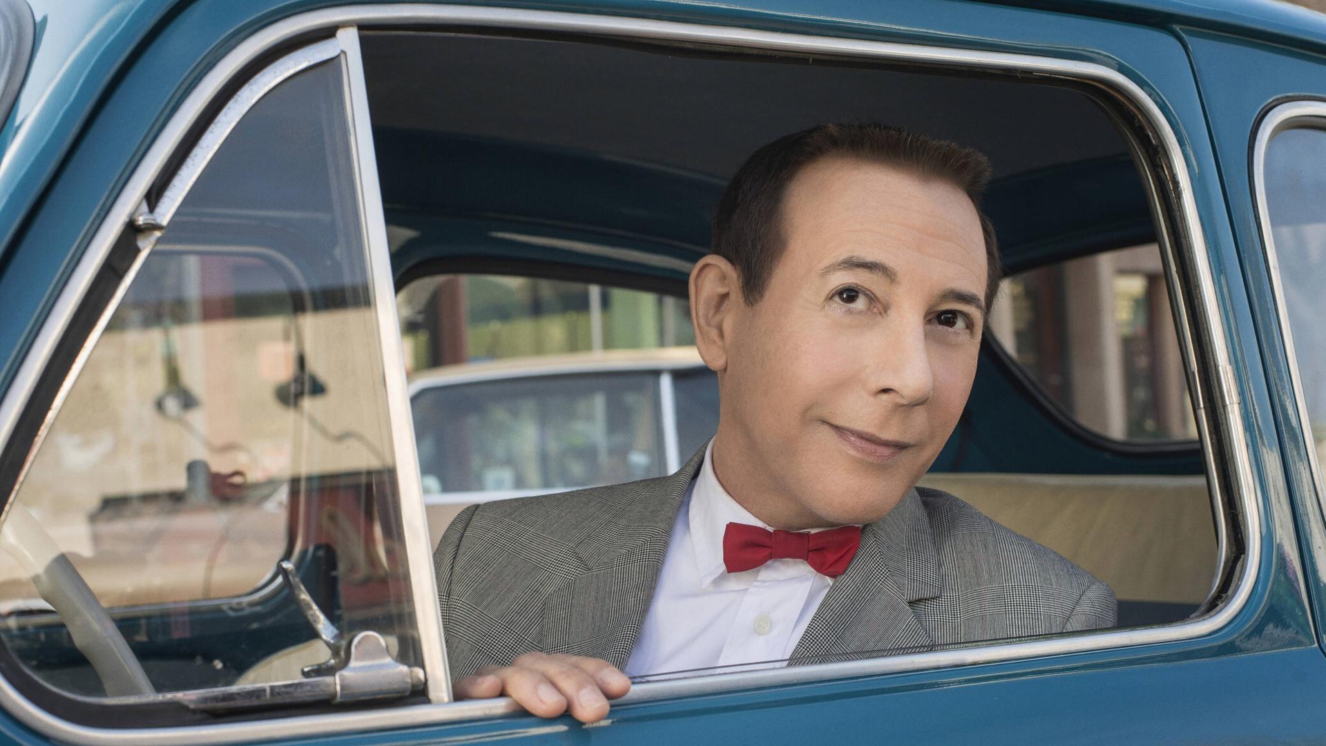 Der Schauspieler Paul Reubens in seiner Rolle als "Pee-Wee Herman". Er schaut aus einem Autofenster und trägt eine rote Fliege.