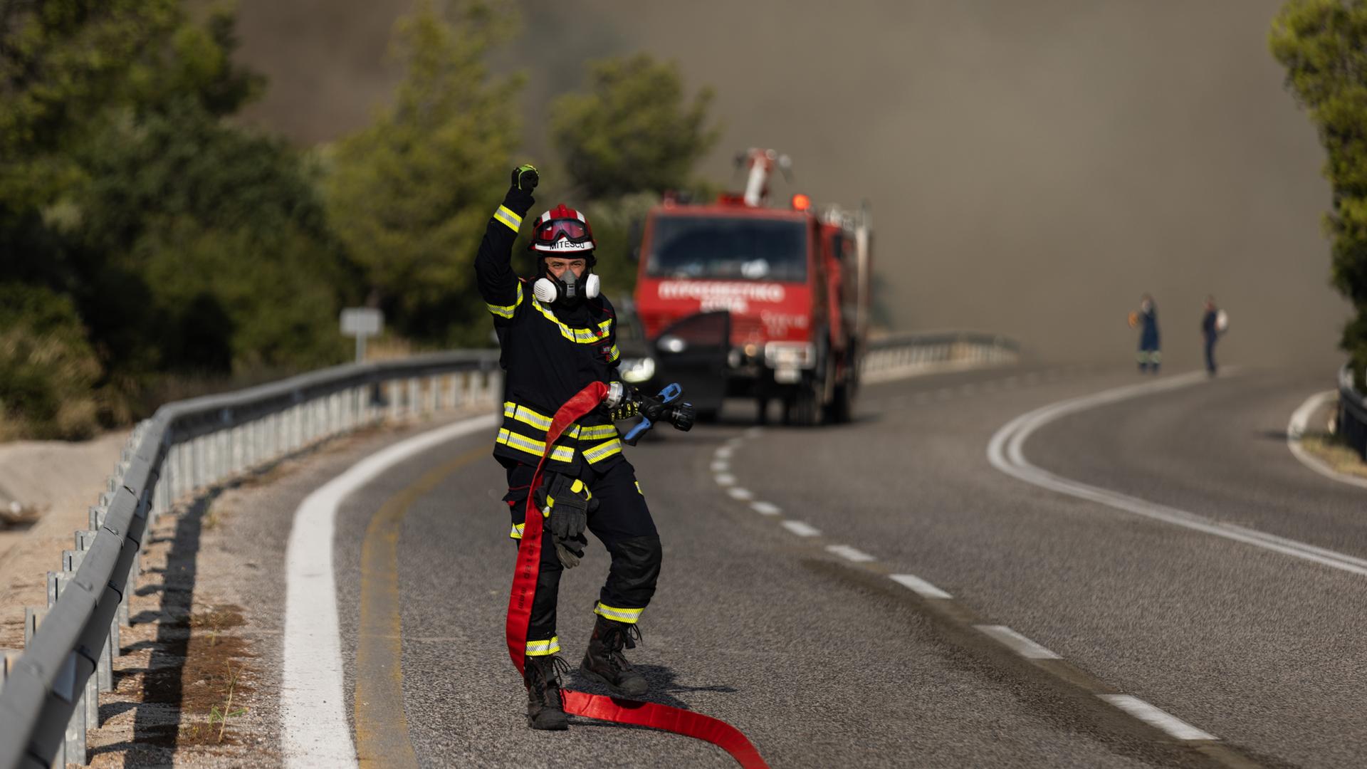 Ein Feuerwehrmann steht im einem Schlauch und Atemschutzgerät auf einer Straße, im Hintergrund raucht es stark aus einem Waldstück.