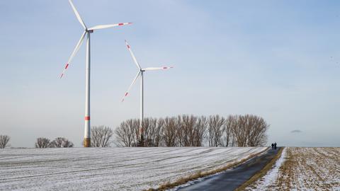 08.02.2019, Bayern, Sindersdorf: Spaziergänger gehen oberhalb eines mit Schnee bedeckten Ackers an Windrädern vorbei. 