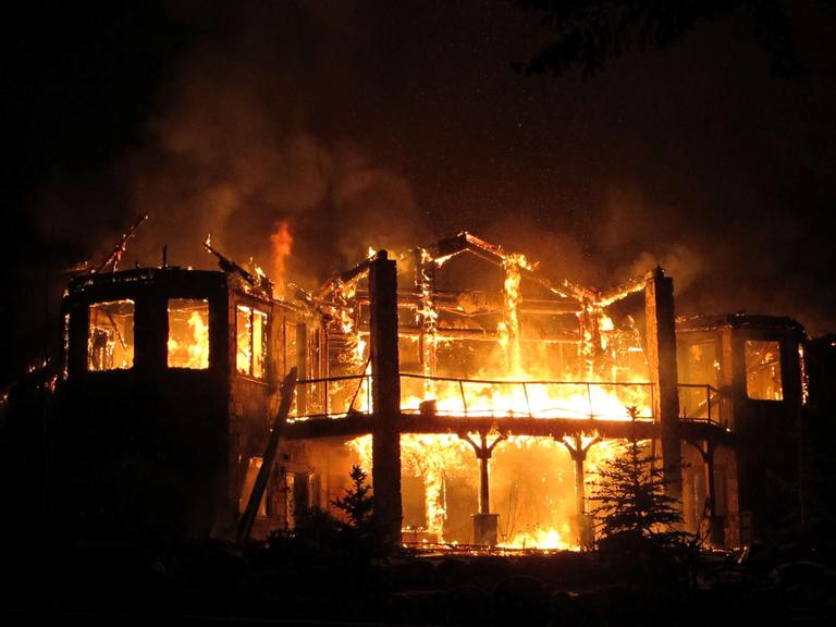 Ein Haus steht bei Nacht vollständig in Flammen, nur die Struktur des Hauses ist noch zu erkennen.