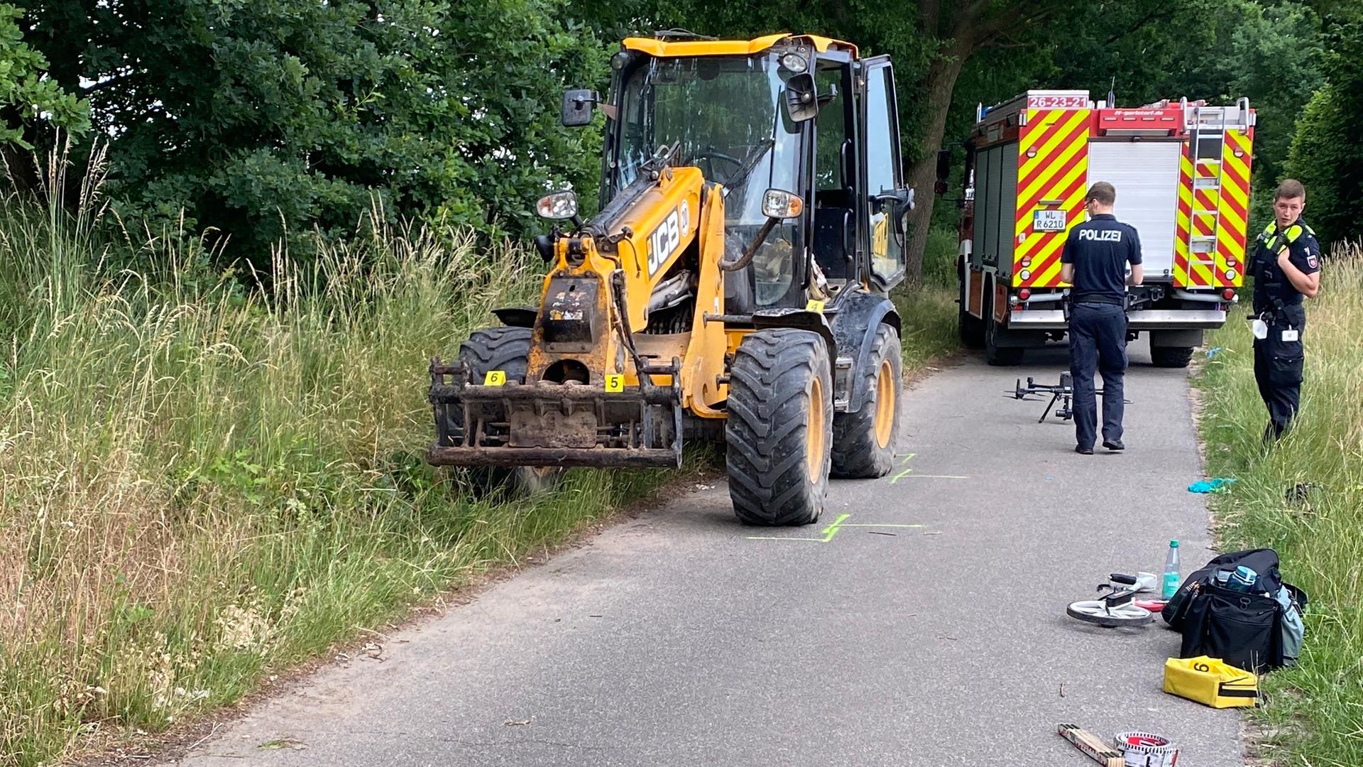 Der Bagger steht am Rande eines asphaltierten Feldwegs an einem Waldrand. Dahinter ein Fahrzeug der Feuerwehr. Zwei Polizisten stehen daneben. Der Bagger steht am Rande eines asphaltierten Feldwegs an einem Waldrand. Dahinter ein Fahrzeug der Feuerwehr. Zwei Polizisten stehen daneben.