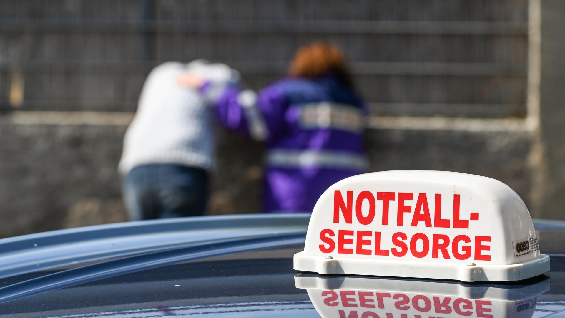 Auf dem Dach eines Autos ist das Schild der Notfallseelsorge zu sehen. Im Hintergrund spricht eine Notfallseelsorgerin mit einem Mann.