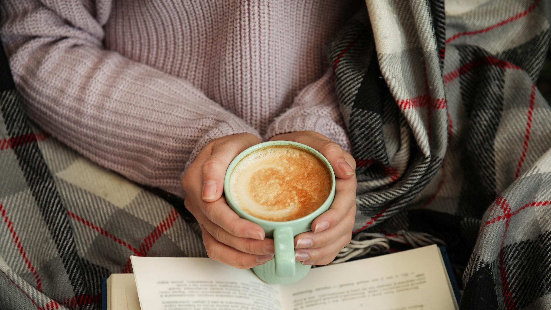 Hände einer weiblich gelesenen Person, die eine Tasse Cappuccino halten, über einem Buch. Die Person, zu der die Hände gehören, scheint in einer Decke eingewickelt zu sein und gemütlich zu lesen.