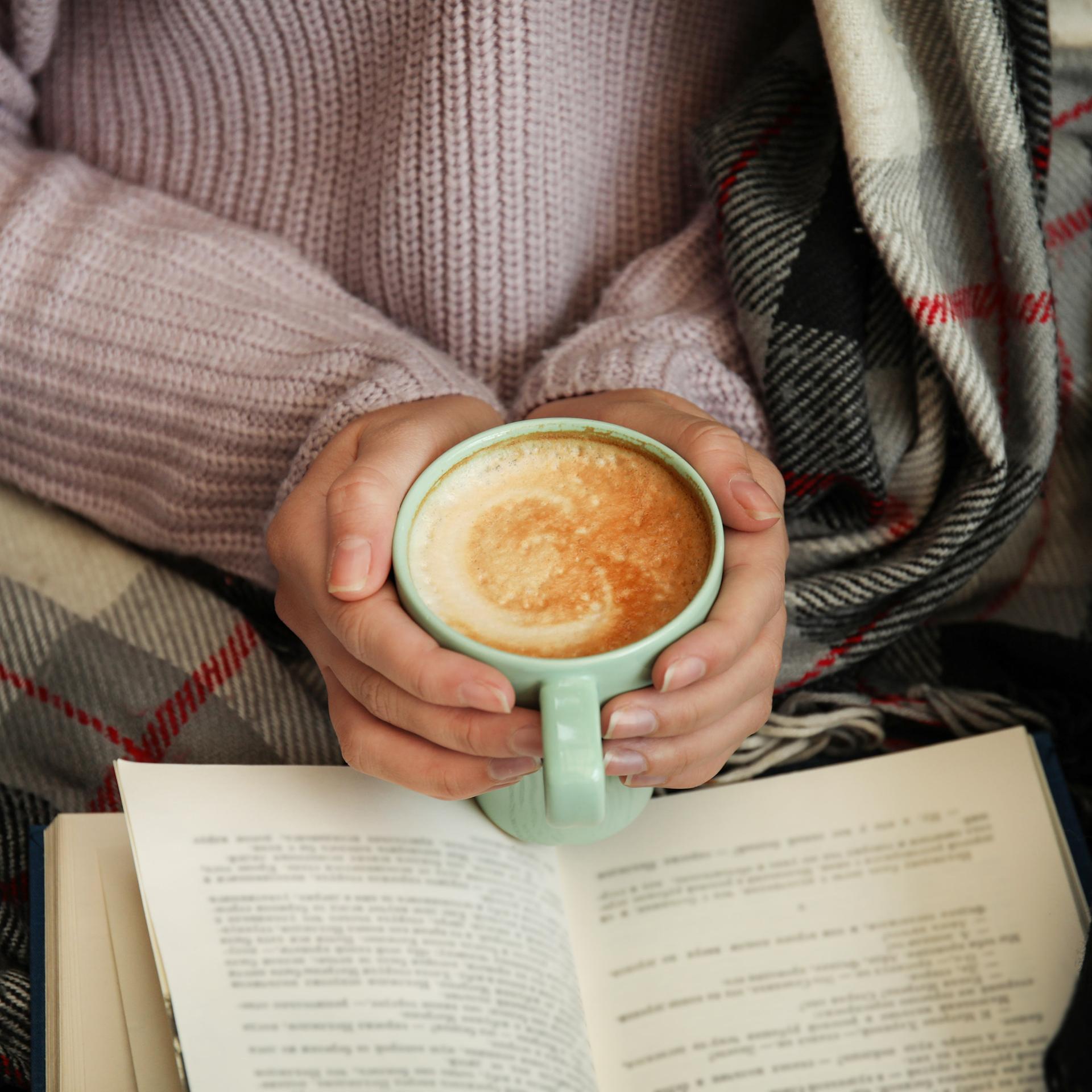 Hände einer weiblich gelesenen Person, die eine Tasse Cappuccino halten, über einem Buch. Die Person, zu der die Hände gehören, scheint in einer Decke eingewickelt zu sein und gemütlich zu lesen.