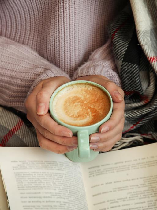 Hände einer weiblich gelesenen Person, die eine Tasse Cappuccino halten, über einem Buch. Die Person, zu der die Hände gehören, scheint in einer Decke eingewickelt zu sein und gemütlich zu lesen.