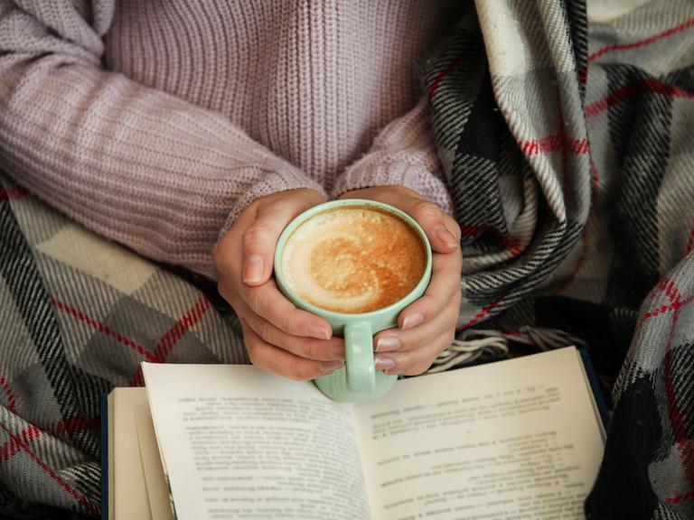 Hände einer weiblich gelesenen Person, die eine Tasse Cappuccino halten, über einem Buch. Die Person, zu der die Hände gehören, scheint in einer Decke eingewickelt zu sein und gemütlich zu lesen.