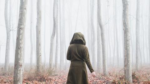 Von hinten ist eine Frau im Kapuzenmantel zu sehen, die durch einen Wald läuft.