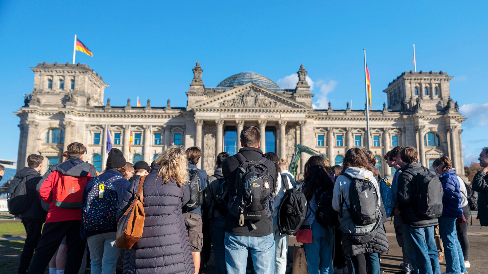 Vor dem Reichstagsgebäude steht im Vordergrund eine Gruppe junger Menschen in Rückenansicht, die auf das Gebäude blicken; die Sonne scheint (19.10.2022)