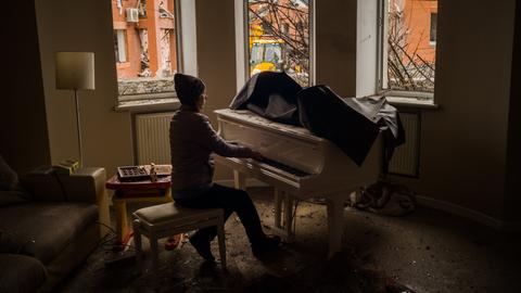 Eine junge Frau spielt Klavier. Durch die zersprungenen Fenster ihrer Wohnung sind im Hintergrund zerstörte Gebäude in der Nachbarschaft zu sehen.