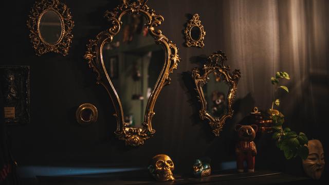 Goldene, verschnörkelte Spiegel in einem dunklen Raum mit düsterer Anmutung. Ein goldener Totenkopf liegt auf einer Ablage vor den Spiegeln.