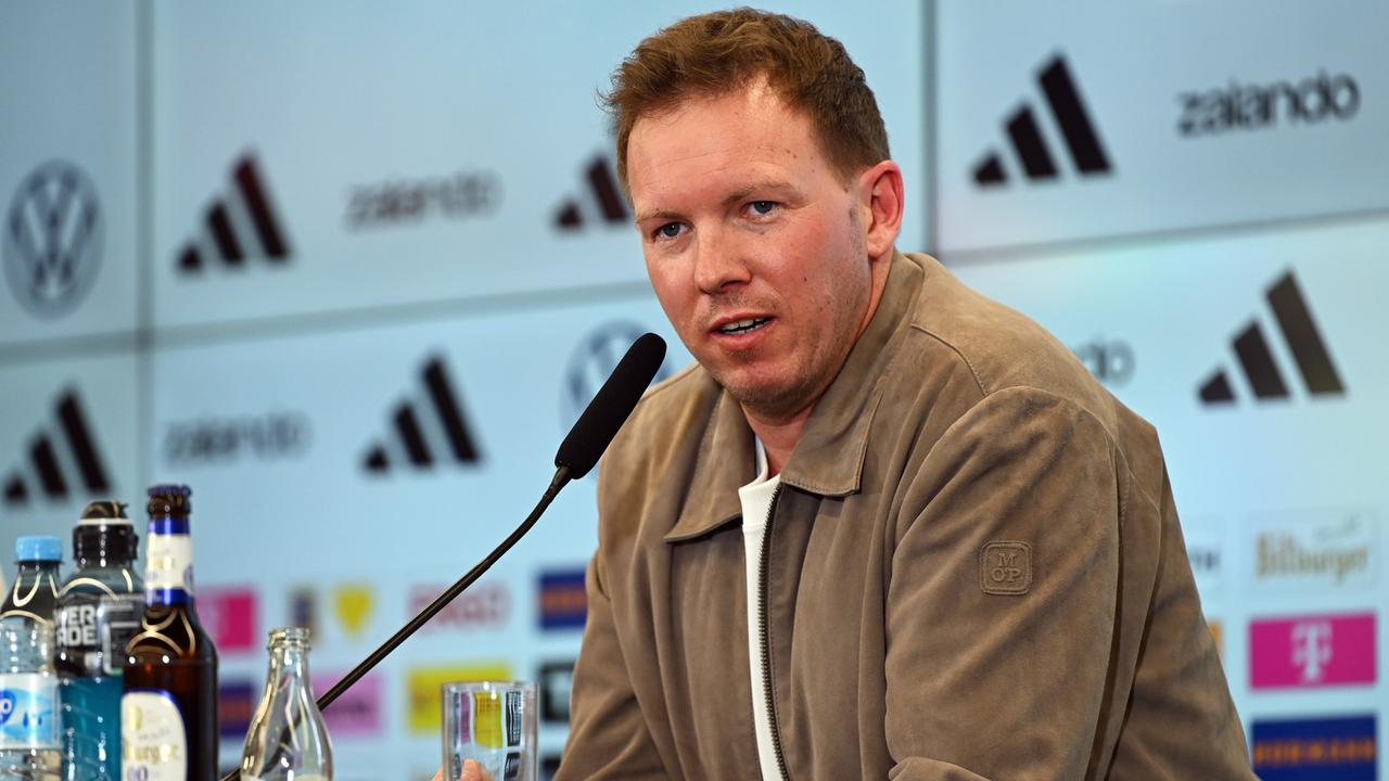 Das Bild zeigt Julian Nagelsmann während einer Pressekonferenz hinter einem Mikrofon. Er trägt ein weißes T-Shirt und darüber eine legere, hellbraune Jacke.