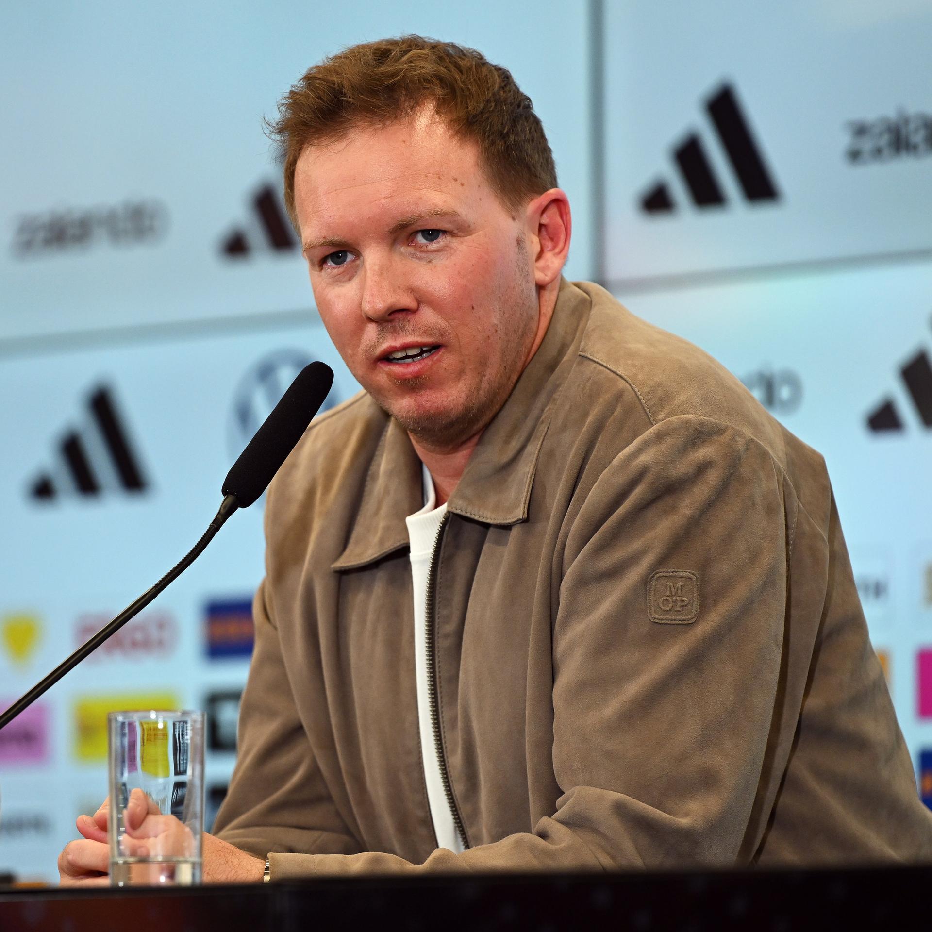 Das Bild zeigt Julian Nagelsmann während einer Pressekonferenz hinter einem Mikrofon. Er trägt ein weißes T-Shirt und darüber eine legere, hellbraune Jacke.